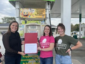 Three women by gas pumps holding a certificate