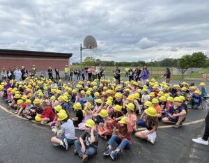 A large group of children sitting on the ground wearing hardhats