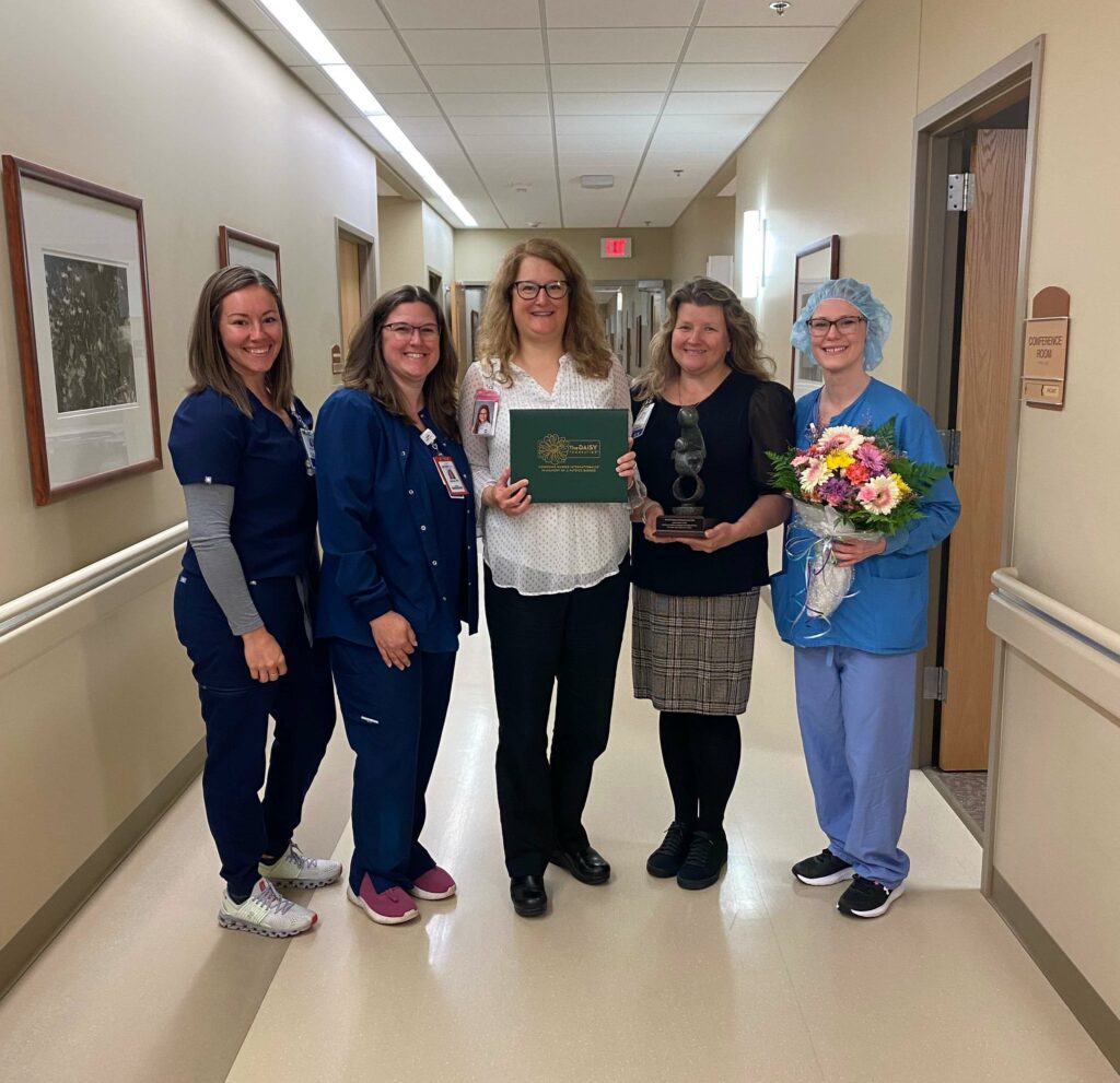 Five women in a hospital hallway with a certificate and flowers