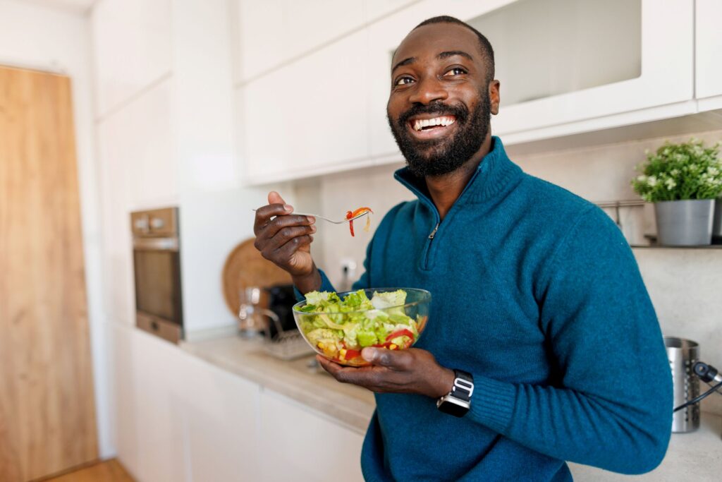 A smiling man eating a salad in a kitchen