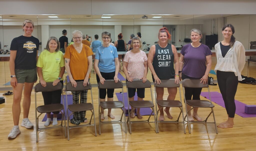 A line of eight people standing behind chairs in a gym