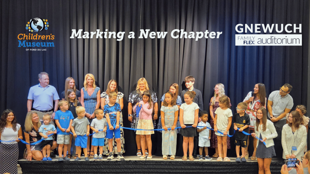 A large group of children and a couple of adults on stage for a ribbon cutting.