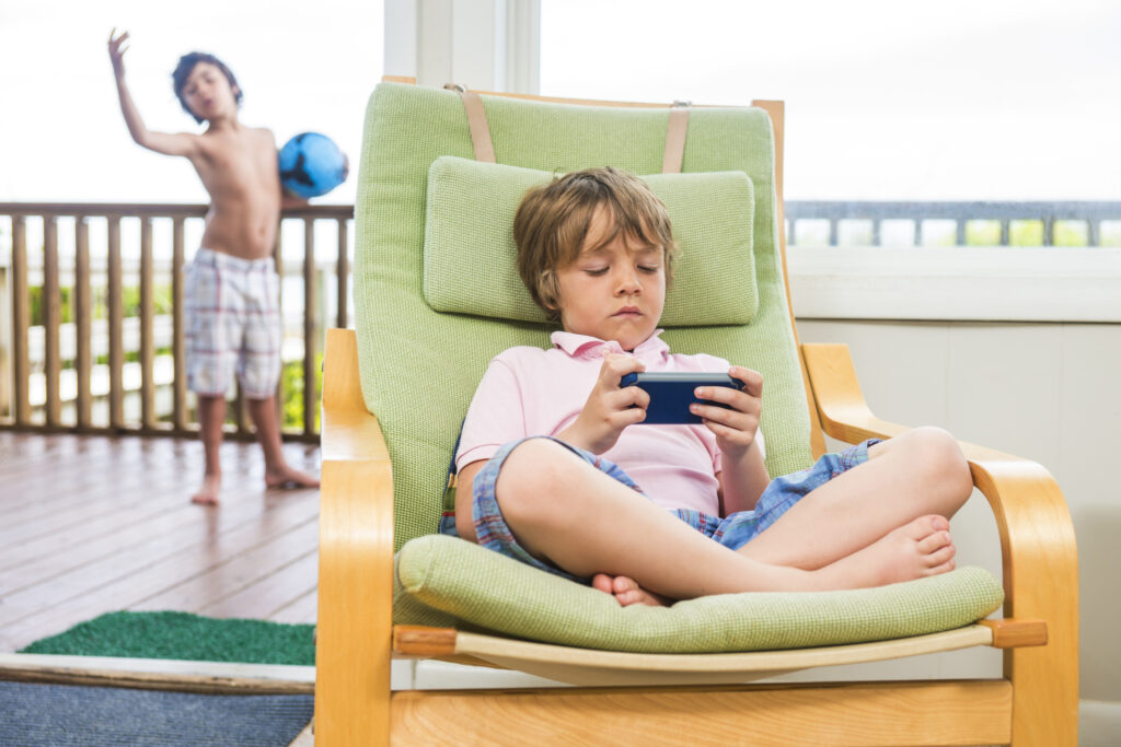 Two children outside. One with a ball and one with a videogame system.